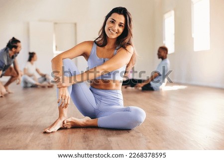 Fit young woman smiling at the camera while sitting in a yoga studio with her class in the background. Happy young woman having a workout session with her class in a fitness studio.