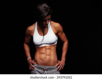 Fit young woman in sportswear looking down while standing with her hands on her hips. Muscular female model posing against black background.