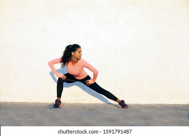 Fit Young Woman Stretching And Doing Squats On The White Concrete Wall Background, Looking To The Right, Wearing Black Sport Tights And Pink Long Sleeve Shirt