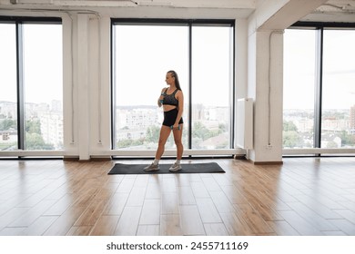 Fit young woman takes break during an intense workout in spacious modern gym with large windows overlooking city. Sports woman stands on yoga mat, demonstrating commitment to fitness and health. - Powered by Shutterstock