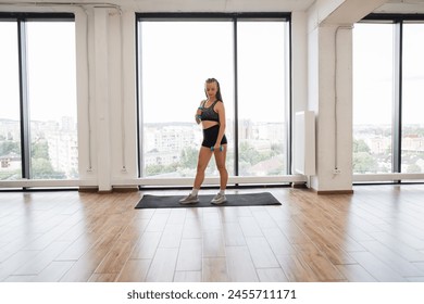 Fit young woman takes break during an intense workout in spacious modern gym with large windows overlooking city. Sports woman stands on yoga mat, demonstrating commitment to fitness and health. - Powered by Shutterstock