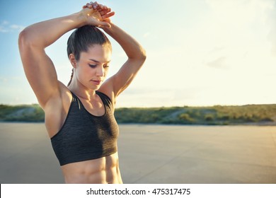 Fit young woman with toned abdominal muscles doing warming up exercises before a workout raising her arms above her head, rural road with copy space - Powered by Shutterstock