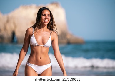 Fit Young Woman In White Swimwear Walking In Wavy Sea And Looking Away While Spending Summer Vacation On Beach In Algarve