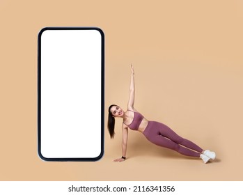 Fitness App. Cheerful Athletic Female Doing Side Plank And Raising Arm Exercising Near Huge Mobile Phone Wearing Sportswear Over Beige Background. Sport Motivation And Online Gym Concept. Mockup