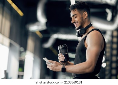 Fitness App. Handsome Smiling Arab Sporty Man Using Smartphone While Standing In Gym, Happy Middle Eastern Male Athlete Browsing New Mobile Application For Workout Productivity Tracking, Copy Space - Powered by Shutterstock