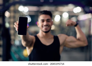 Fitness App. Sporty Muscular Arab Man Holding Smartphone With Blank Black Screen And Showing Biceps At Camera, Athlete Guy Recommending Workout Application While Standing In Modern Gym, Mockup