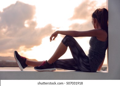 Fitness athlete sports woman relaxing after workout sitting on window looking at sunset view. Silhouette of runner in activewear resting at home on outdoor terrace in sky background. - Powered by Shutterstock