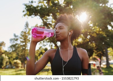 Fitness athlete young african american woman listening to music on earphones drinking water in a reusable water bottle after working out exercising on sunny day at the park
