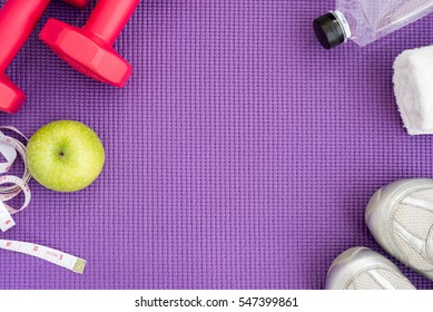 Fitness background with red dumbbells, green apple, sport shoes, white towel, water and measuring tape over violet yoga mat with copy space. Healthy and lifestyle concept
