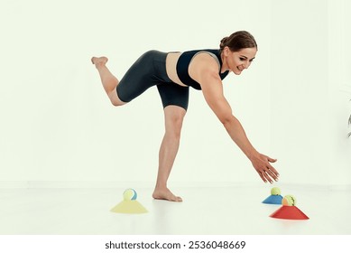 A fitness coach performs a dynamic balance exercise, reaching towards colorful cones with tennis balls. This image highlights coordination, balance, and core strength training.. - Powered by Shutterstock