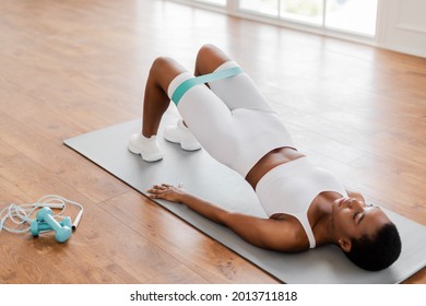 Fitness Concept. Athletic Black Woman In White Sportswear Training With Elastic Resistance Band Above Knees In Fitness Studio Or Living Room. Lady Doing Glute Bridge Exercise For Buttocks And Legs