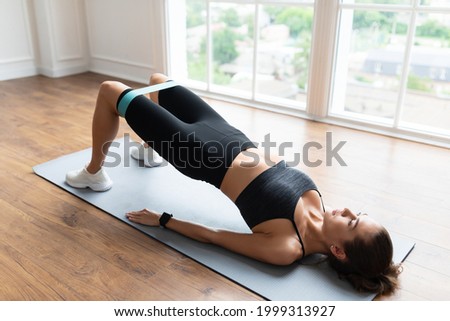 Fitness Concept. Athletic woman in black sportwear training with elastic resistance band above knees in fitness studio or in living room. Lady doing glute bridge exercise for buttocks, legs and core
