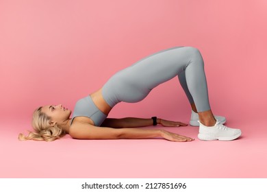 Fitness Concept. Athletic young woman in gray sportswear doing glute bridge exercise for buttocks and legs muscles. Focused lady training isolated over pink studio background, profile lide view - Powered by Shutterstock