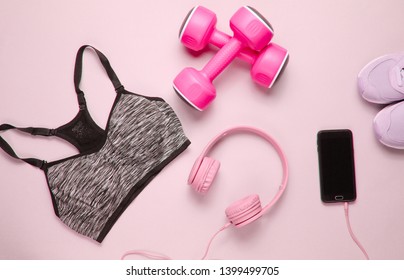 Fitness concept. Flat lay of a smartphone with headphones, plastic dumbbells, sports bra, sneakers on pink background. Top view