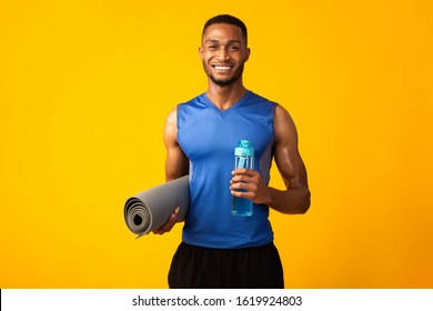 Fitness Concept. Happy Black Man Holding Water Bottle And Yoga Mat, Smiling At Camera Over Yellow Wall, Copyspace