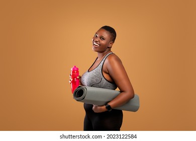 Fitness Concept. Portrait of happy black plus size woman holding water bottle and yoga mat, body positive female smiling and looking at camera standing isolated over brown studio background