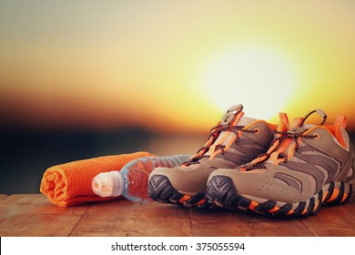 Fitness Concept With Sport Footwear, Towel And Water Bottle Over Wooden Table In Front Of Sunset Landscape. 