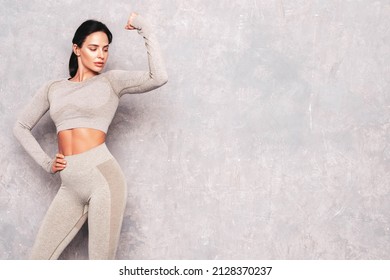 Fitness Confident Woman In Sports Clothing. Sexy Young Beautiful Model With Perfect Body. Female Posing Near Grey Wall In Studio. Stretching Out Before Training.Shows Her Muscles