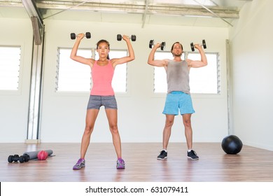 Fitness Couple Working Out Together Training Arms Doing Overhead Shoulder Press With Dumbbells Weights Or Standing Dumbbell Press Exercise At Gym. Asian Woman And Caucasian Man.