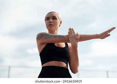 Fitness enthusiast performs arm stretches, focusing on her pre-workout routine against a cloudy sky - Powered by Shutterstock