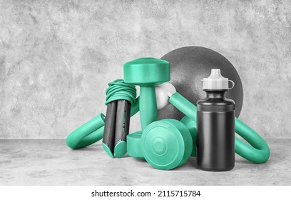 Fitness equipment on a gray concrete background with copy space. Dumbbells, sport bottle of water, jump rope, leg trainer, gymnastic ball, front view. Home workout. Fitness and activity.