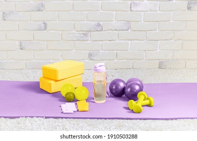 Fitness equipment on a yoga mat on a brick wall background. A bottle of water, rubber bands, yoga blocks, balls and dumbbells. Exercise equipment for home workout. Selective focus. Copy space. - Powered by Shutterstock