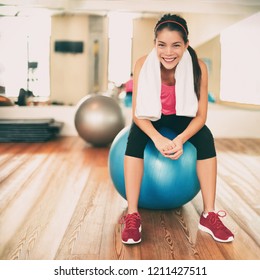 Fitness Girl Exercising Happy In Gym Resting On Pilates Exercise Ball After Training Class. Asian Woman Sweating With Towel On Weight Loss Workout Portrait. Active Healthy Lifestyle.