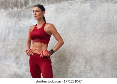 Fitness Girl Portrait. Woman In Fashion Sporty Outfit Standing Against Stone Wall After Workout. Fit Female With Strong Muscular Body Keeping Hands On Hips And Looking Away. 