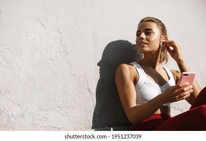 Fitness Girl Sitting Outdoors With Smartphone. Female Athlete Listening Music And Relaxing After Jogging, Workout Outside On Fresh Air, Look Away And Smile, Enjoy Good Workout