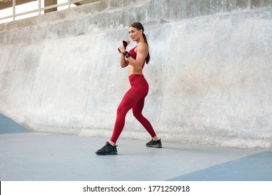 Fitness Girl Stands In Boxing Pose And Getting Ready To Do Kick Workout. Fit Woman With Strong Muscular Body In Fashion Sporty Outfit Exercising At Outdoor Stadium Against Concrete Wall. 