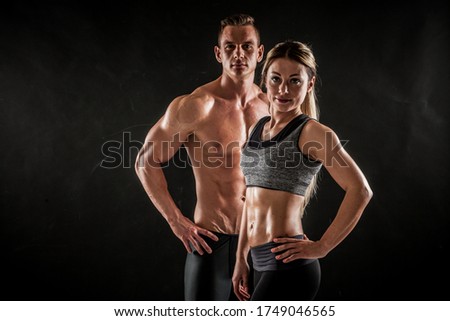 Fitness in gym, sport and healthy lifestyle concept. Couple of athletic man and woman showing their trained bodies on dark background. Two bodybuilder models standing and demonstrating tight muscles.