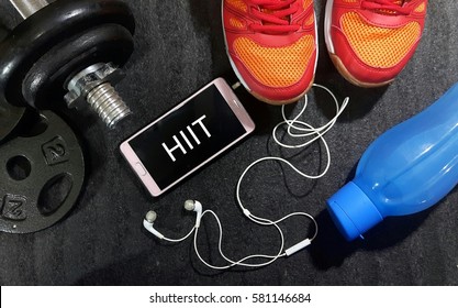 Fitness, Healthy And Active Lifestyles Concept. Dumbbells, Sport Shoes, Smart Phone With Earphone And Water Bottle Gym Floor With Word Hiit