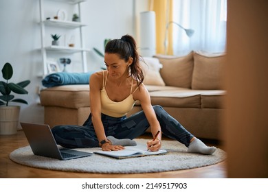 Fitness Instructor Making Exercise Plan And Taking Notes While Using Laptop In The Living Room.