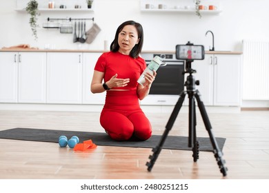 Fitness instructor recording online workout tutorial at home holding water bottle. Showing exercise equipment such as dumbbells and resistance bands. - Powered by Shutterstock