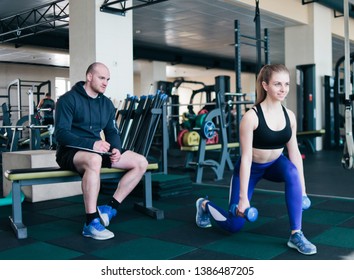 Fitness instructor supervises and notes in a notebook the results of training young athletic blonde performing exercises lunges with dumbbells in her hands in the gym