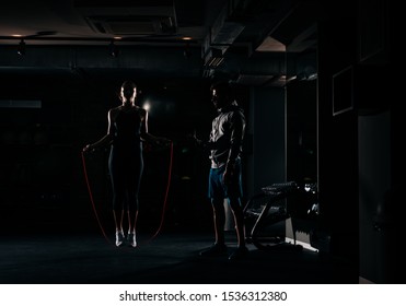 Fitness instructor supervising the female athlete in a gym exercises with jumping rope, high contrast, low key, dark