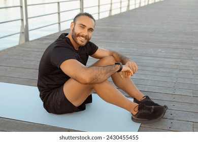 Fitness joy. Happy sporty guy in activewear takes a break on a pier, sitting on gym mat outside, radiating wellbeing and happiness as he smiles at camera. Concept of healthy lifestyle and workout