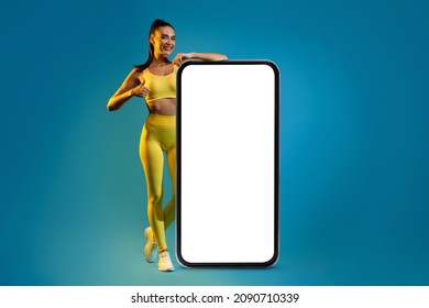 Fitness Lady Leaning On Big Phone With Empty Screen Advertising Mobile Application Gesturing Thumbs Up Standing Over Blue Background, Wearing Yellow Sportswear. Mockup. I Like This Workout App