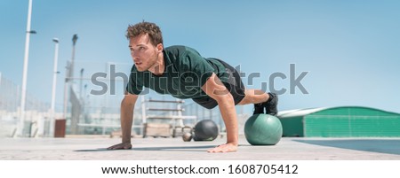 Fitness man banner panorama. Athlete strength training pushup balancing legs on medicine ball for advanced core body workout push-ups floor exercises at outdoor gym.