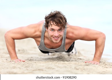 Fitness Man Exercising Push Ups Smiling Happy. Male Fitness Model Cross-training On Beach. Caucasian Man In His Twenties.