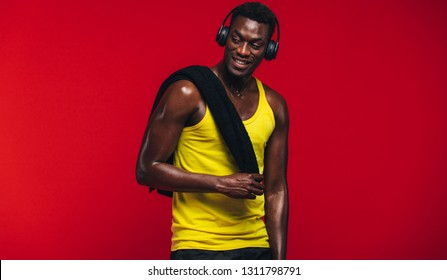 Fitness Man With Towel On Shoulder Listening To Music On Headphones. Fit Young Man Resting After From Workout On Red Background.