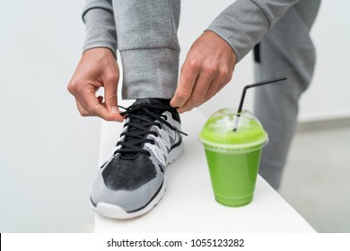 Fitness man tying running shoes and green smoothie breakfast. Health weight loss fitness man drinking green smoothie lacing trainers laces ready for jogging active lifestyle. Morning active lifestyle.