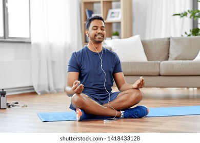 fitness, meditation and healthy lifestyle concept - indian man in earphones listening to music on smartphone and meditating in lotus pose at home - Powered by Shutterstock