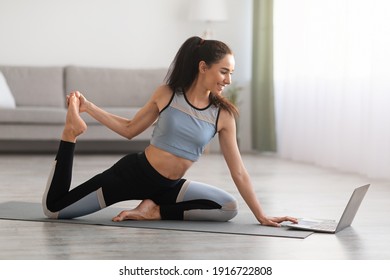 Fitness online. Athletic young woman stretching at living room, sitting on yoga mat in front of laptop, copy space. Good looking brunette lady exercising at home, using video lessons, side view