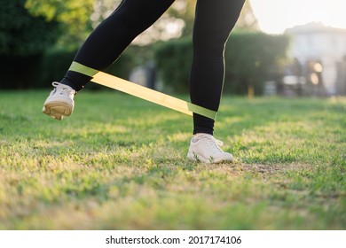 Fitness Outdoors. Morning Exercise. Sport Gear. Healthy Lifestyle. Overweight Woman Doing Legs Workout Outside With Resistance Band On Green Grass Park Landscape.