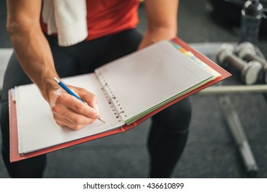 Fitness Personal Trainer Writing On Notebook While Sitting On A Gym Bench. Healthy Sporty Young Man Goals.