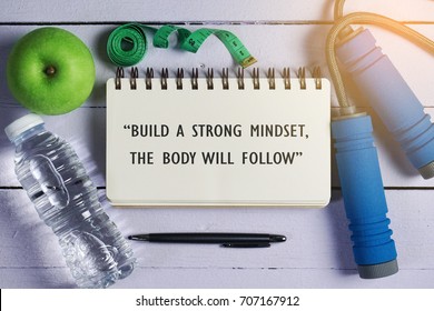 Fitness quotes "Build A Strong Mindset, The Body Will Follow" on open notebook. Water, green apple, measuring tape, jumping rope, pen and open notebook over the wooden background.