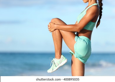 Fitness runner body closeup doing warm-up routine on beach before running, stretching leg muscles with standing single knee to chest stretch. Female athlete preparing legs for cardio workout. - Shutterstock ID 400895035