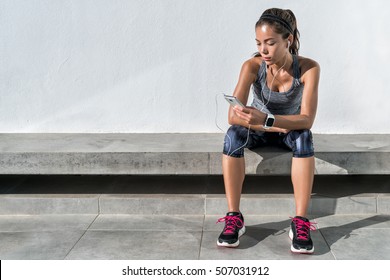 Fitness Runner On Mobile Smart Phone App Tracking Progress Listening To Music With Earphones For Fitness Motivation. Athlete Runner In Sportswear Relaxing Sitting Getting Inspired. Asian Mixed Race.