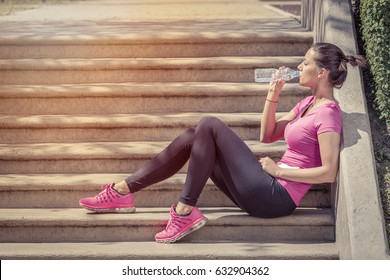 Fitness Runner Woman Drinking Water And Sitting On Staircase. Athlete Girl Taking A Break During Run To Hydrate During Hot Summer Day. Healthy Active Lifestyle.  